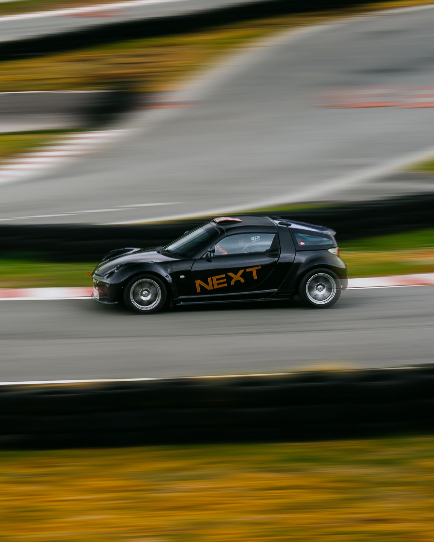 Black sports car with 'NEX1' branding races on a racetrack with motion blur effect and yellow background.