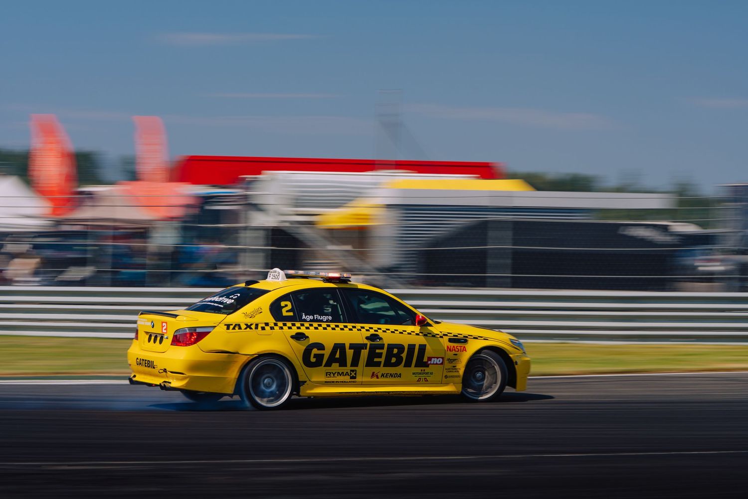Yellow racing car speeds along racetrack with motion blur effect against blue sky background.