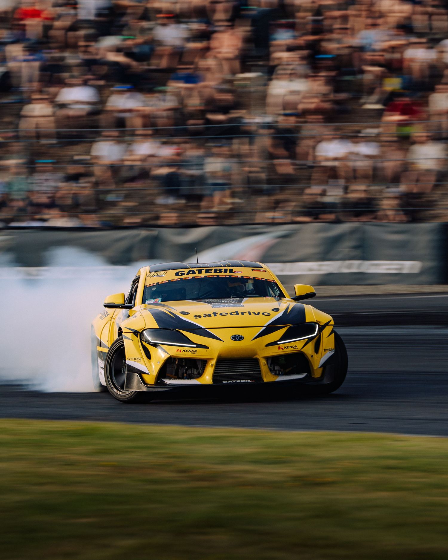 A yellow McLaren race car speeds along a racetrack with motion blur and spectators in the background.