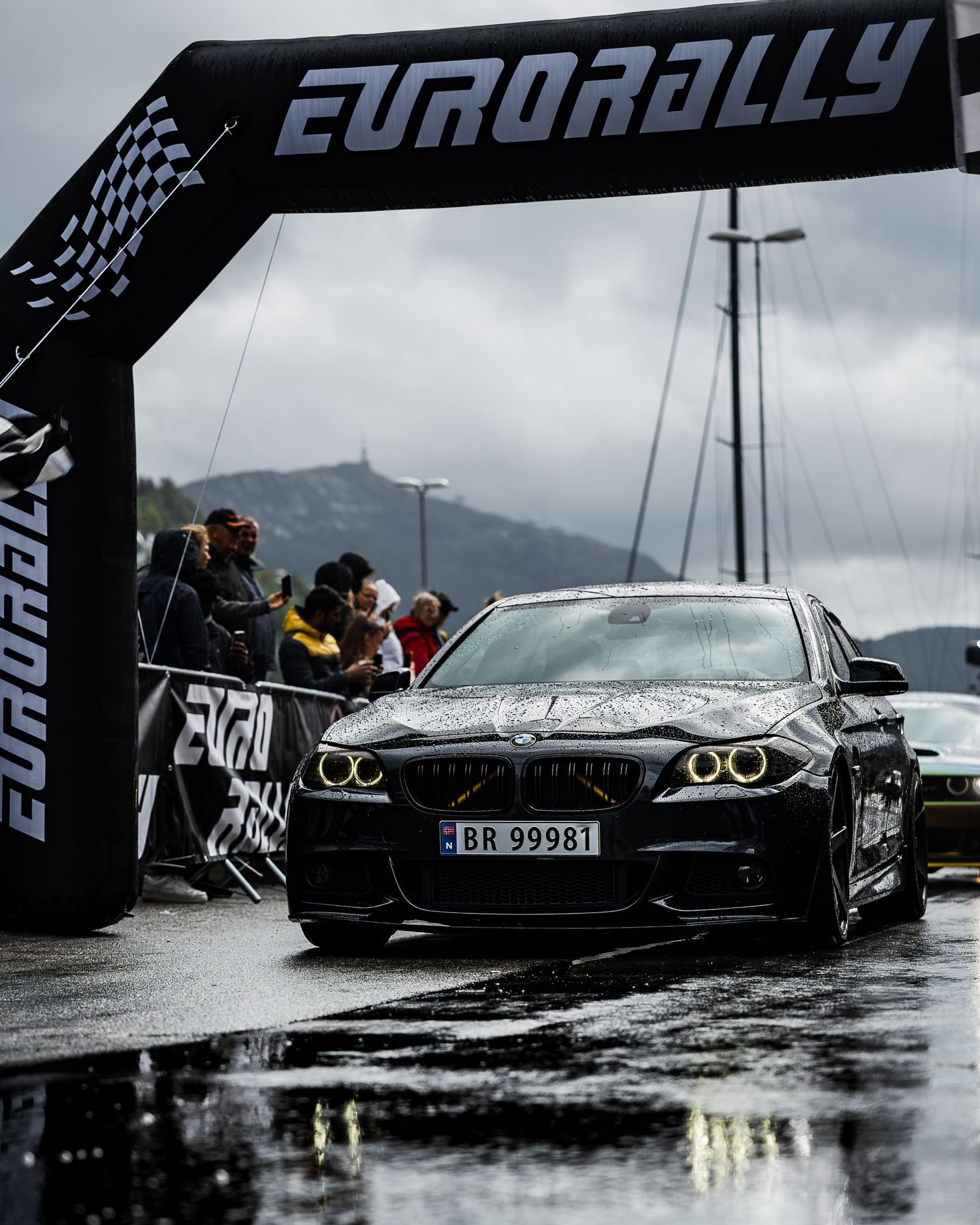 Black BMW M5 crosses finish line during wet Eurorally event with mountain scenery.