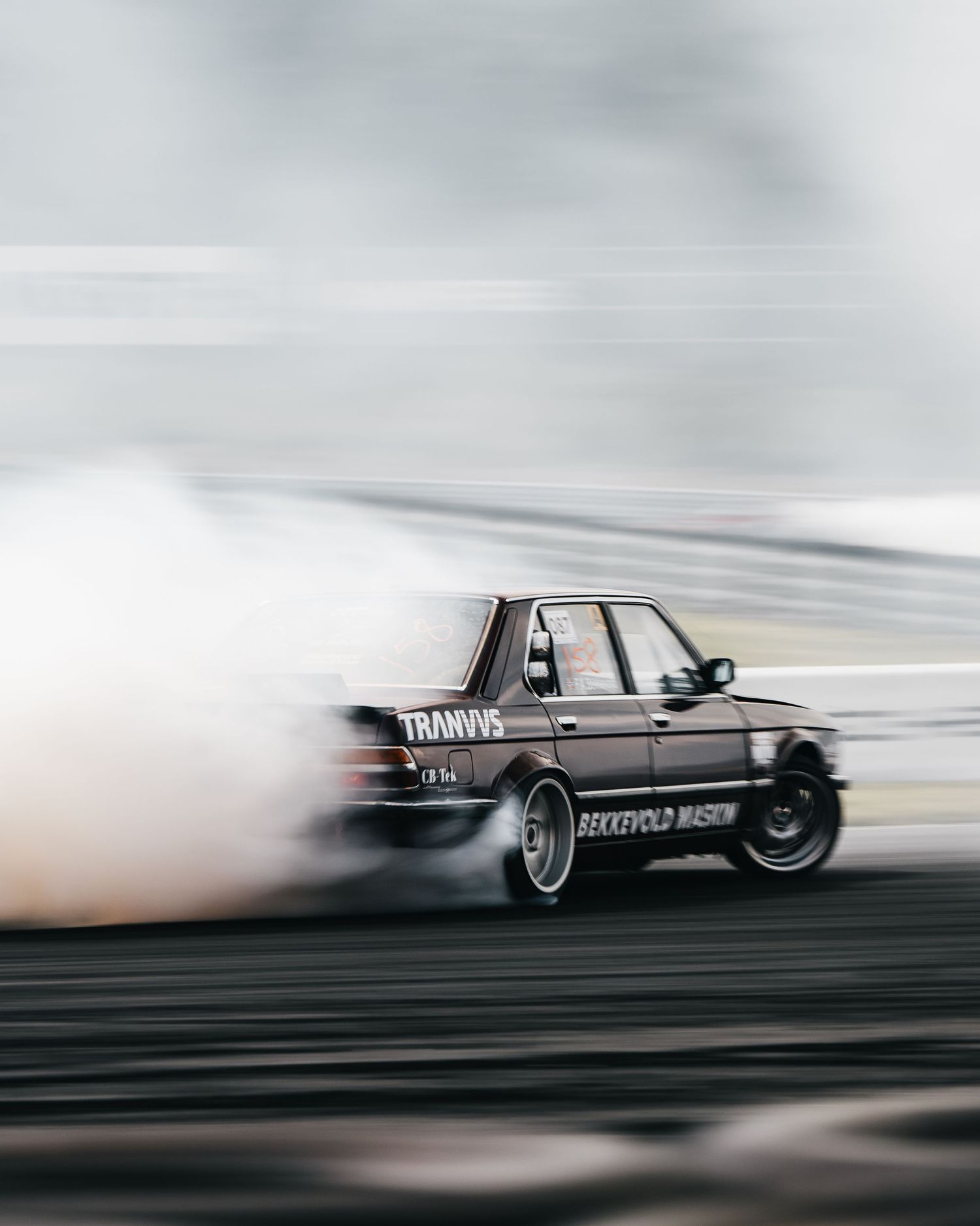 A BMW drift car creates a cloud of tire smoke while drifting sideways on a race track in motion-blurred action.