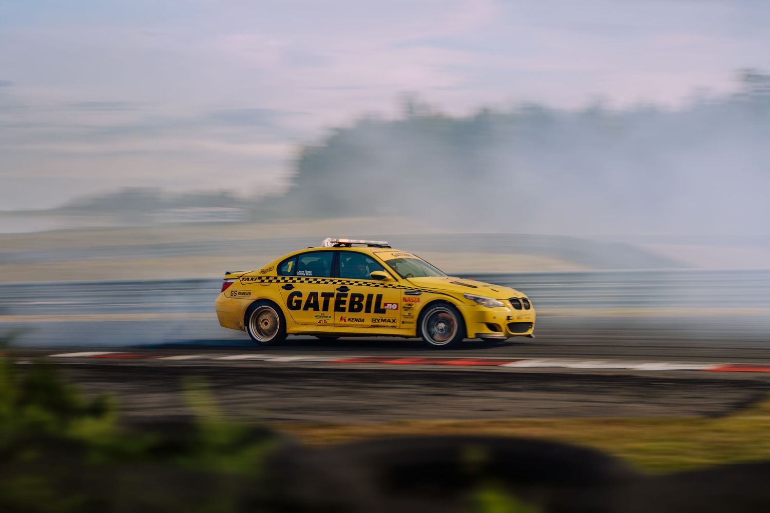 Yellow taxi drifts on a racetrack with smoke billowing from its tires against a cloudy sky at sunset.