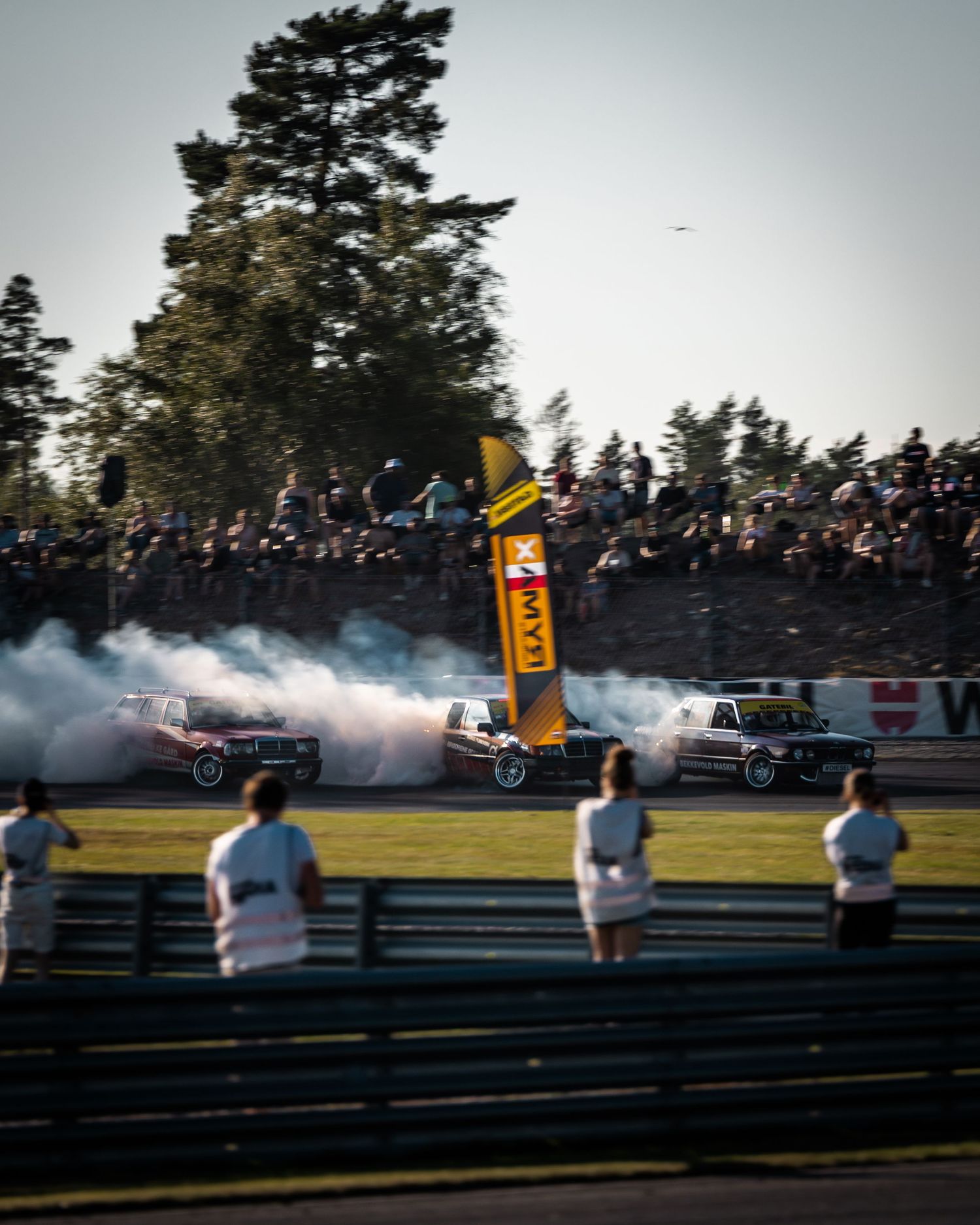 Drift cars perform synchronized burnouts on a race track as spectators watch from behind barriers during a motorsport event.