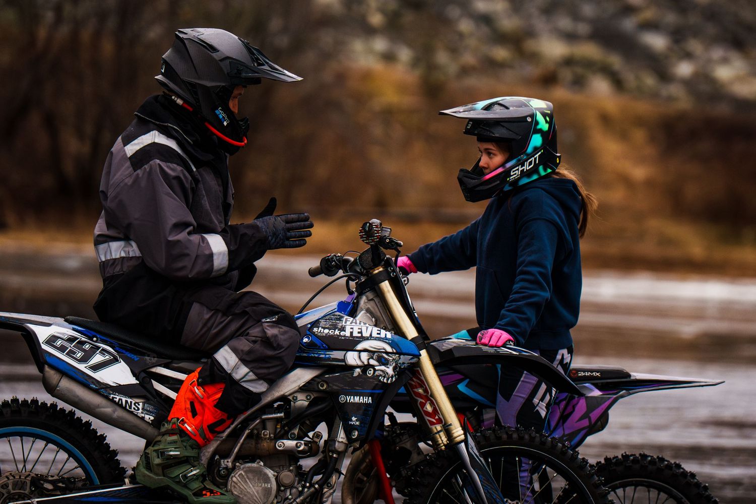 Two dirt bike riders in protective gear having a conversation while stopped on their motorcycles in an outdoor setting.