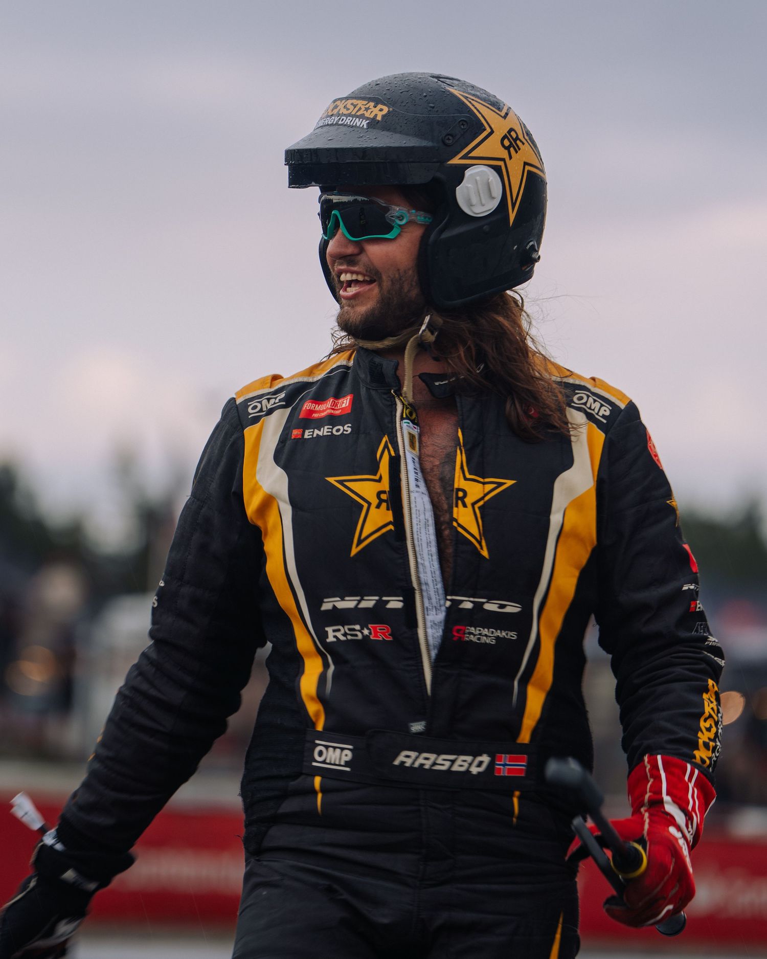 Race car driver wearing black and gold racing suit with sponsor logos and helmet poses at outdoor track.