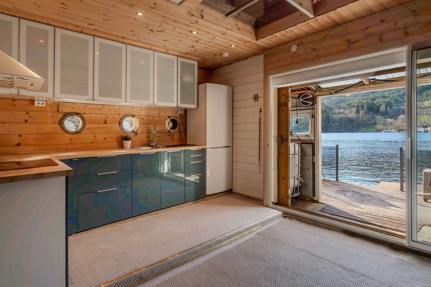 Contemporary kitchen with teal cabinets and wooden walls opening to waterfront view through glass doors.