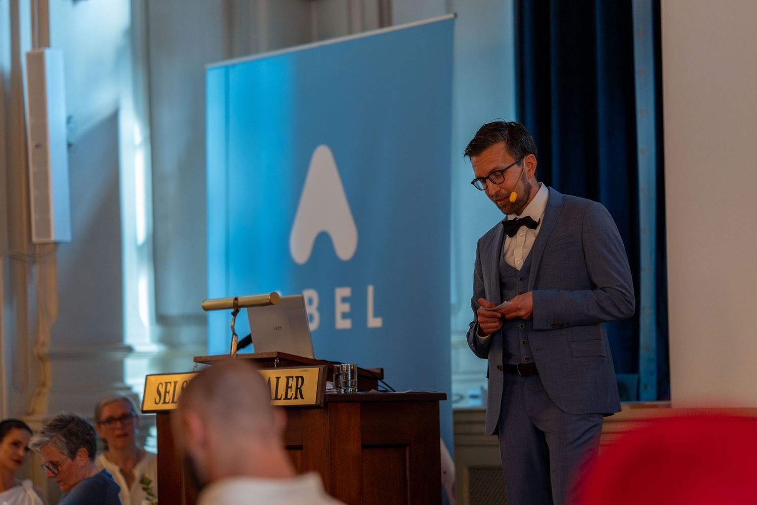 Speaker presents at podium during ABEL corporate event with blue backdrop.