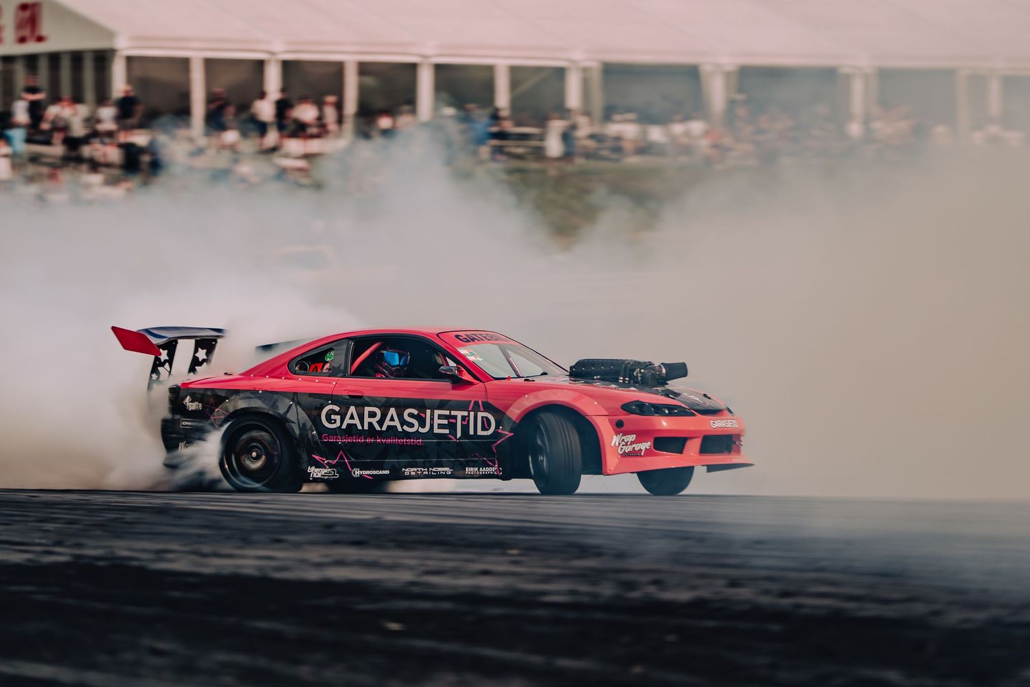 Red drift car performs a high-speed drift maneuver on a race track creating clouds of tire smoke.