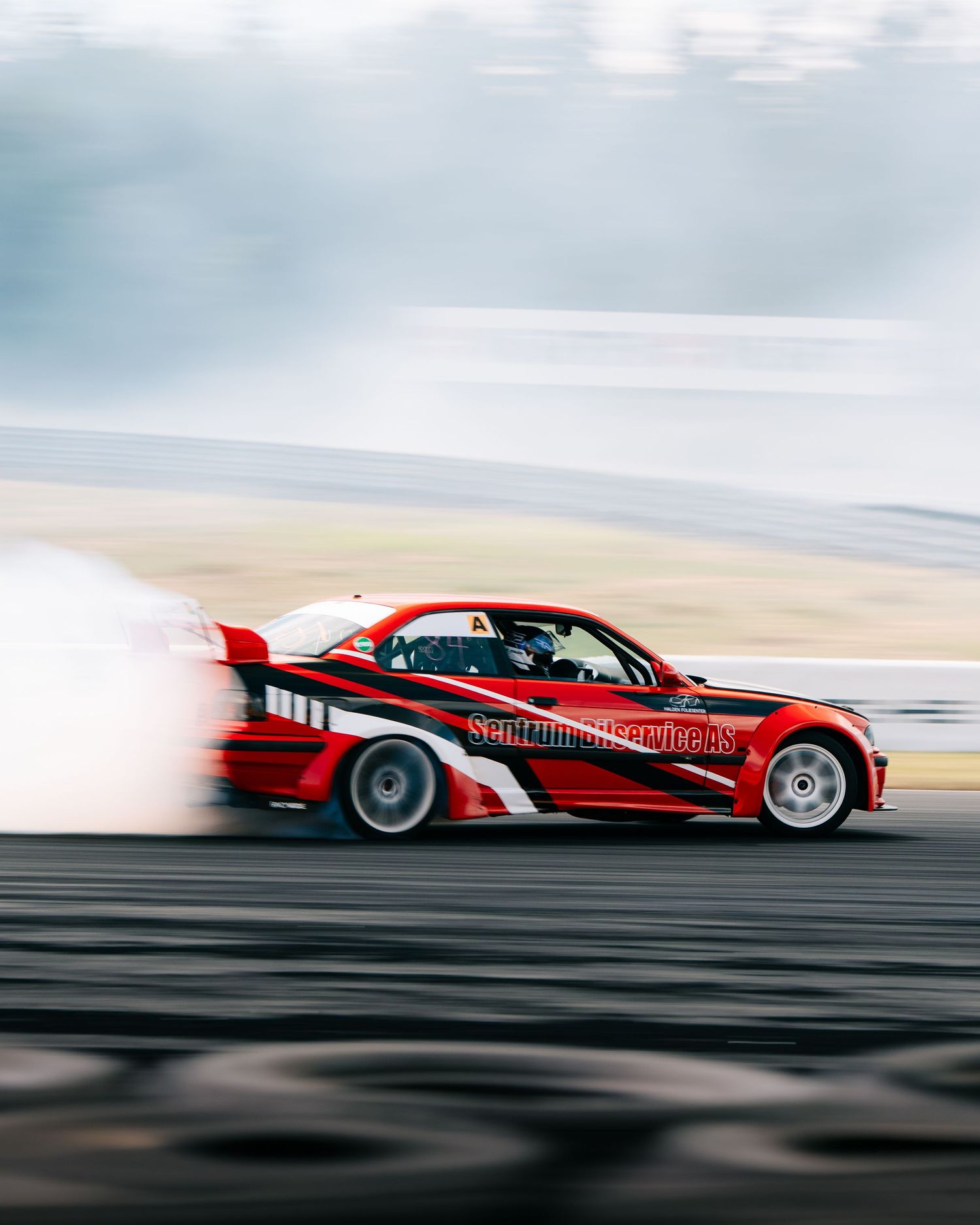 Red drift car with custom graphics performs a smoky drift on a race track during a motorsport competition.