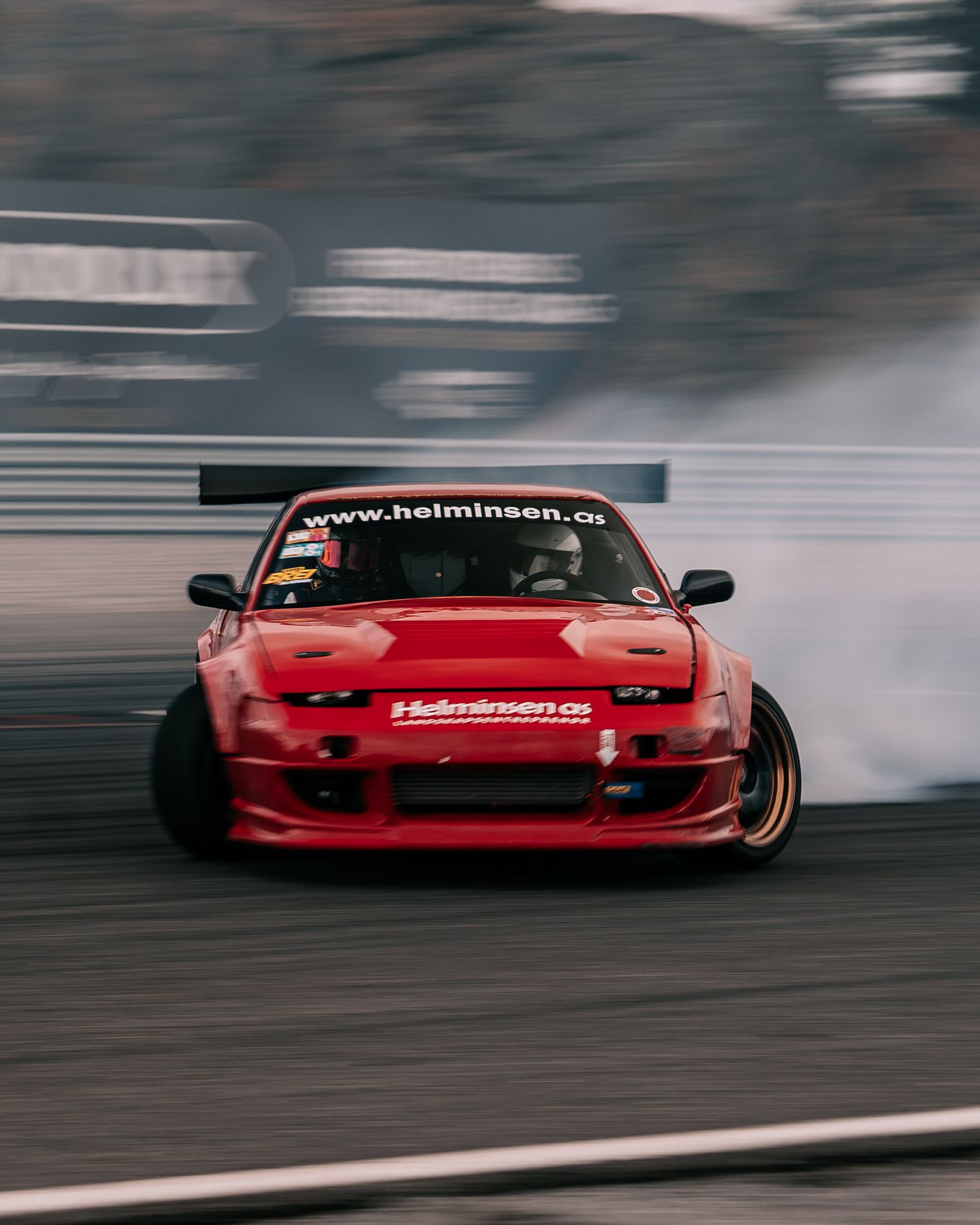 Red drift car slides sideways on track creating smoke while performing controlled drift maneuver.