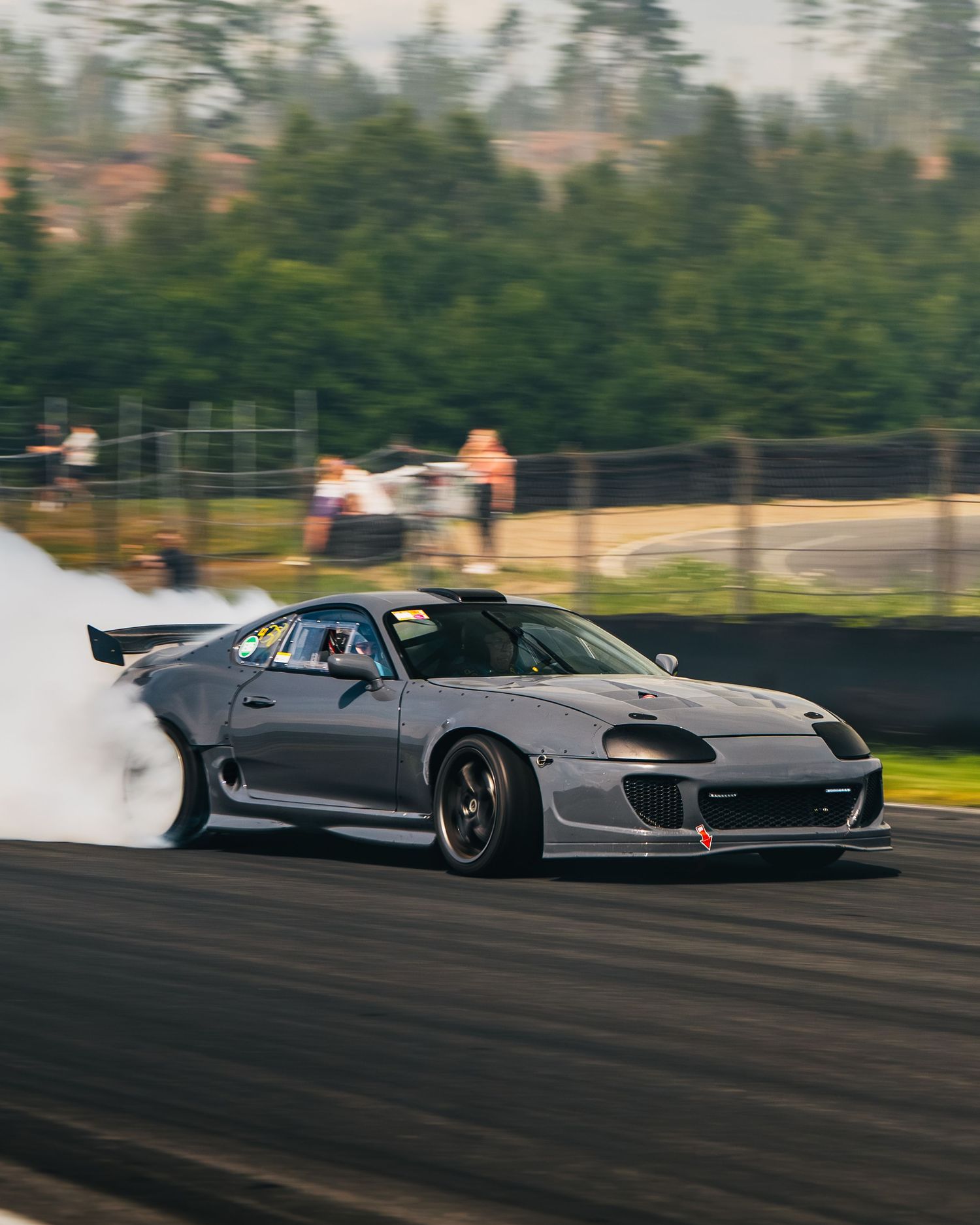 A silver sports car drifts sideways on a race track with smoke billowing from its tires during an intense drift session.