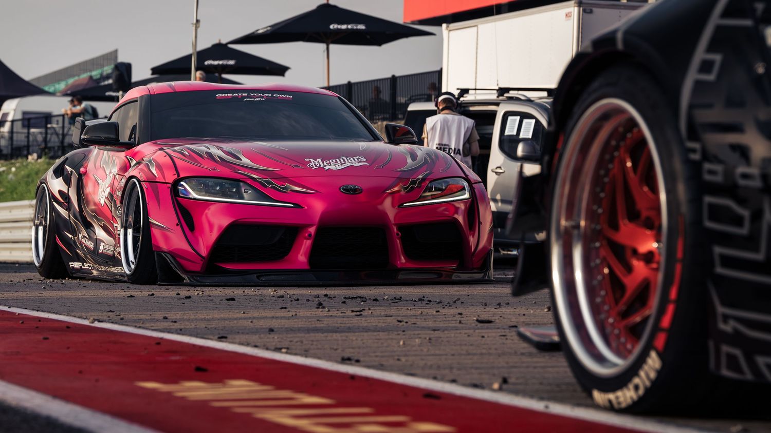 A bright pink Toyota Supra sports car with wide body kit and red wheels parked at a racing event.