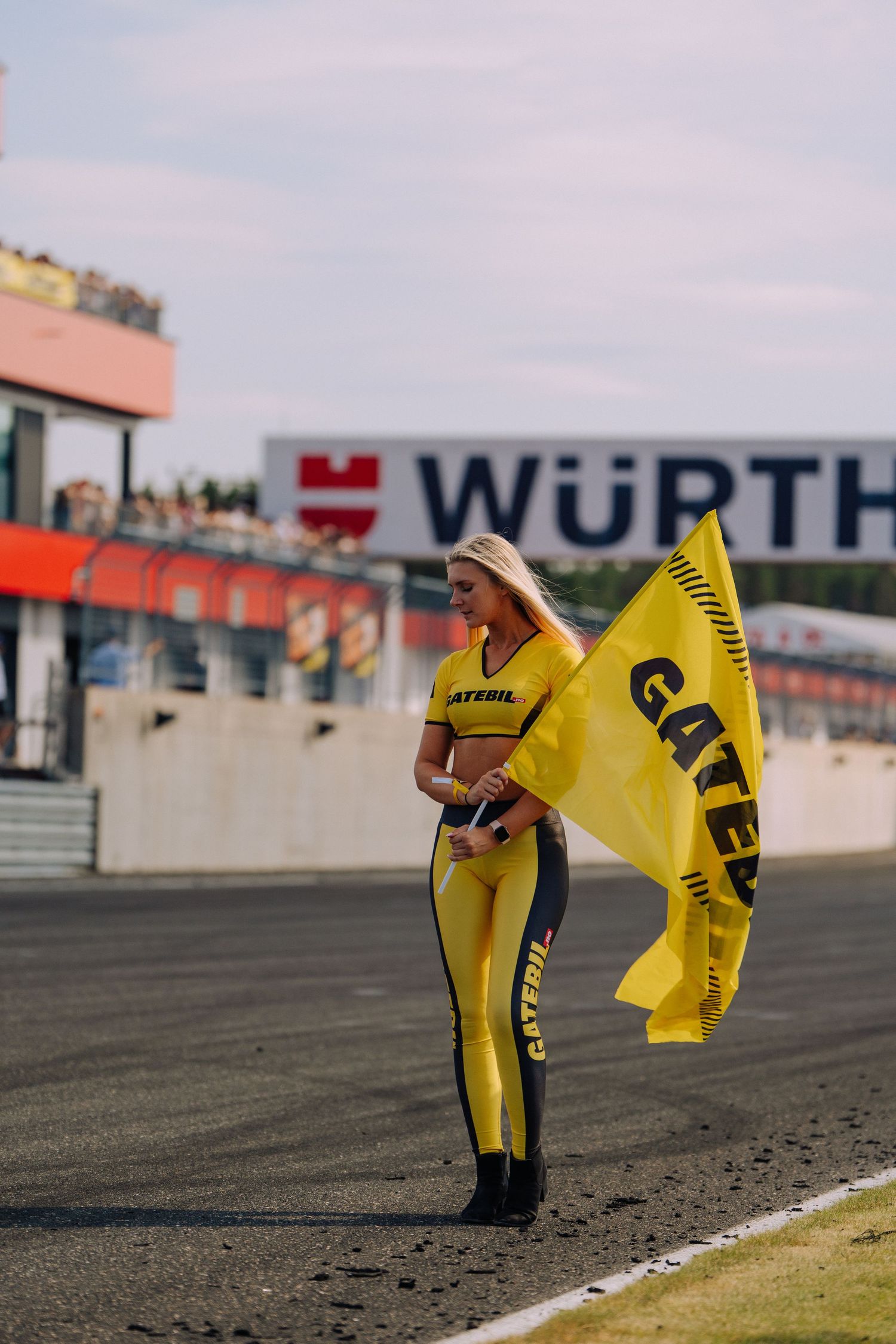 Race track flag bearer in yellow racing suit holds bright yellow caution flag at Wurth motorsport circuit.