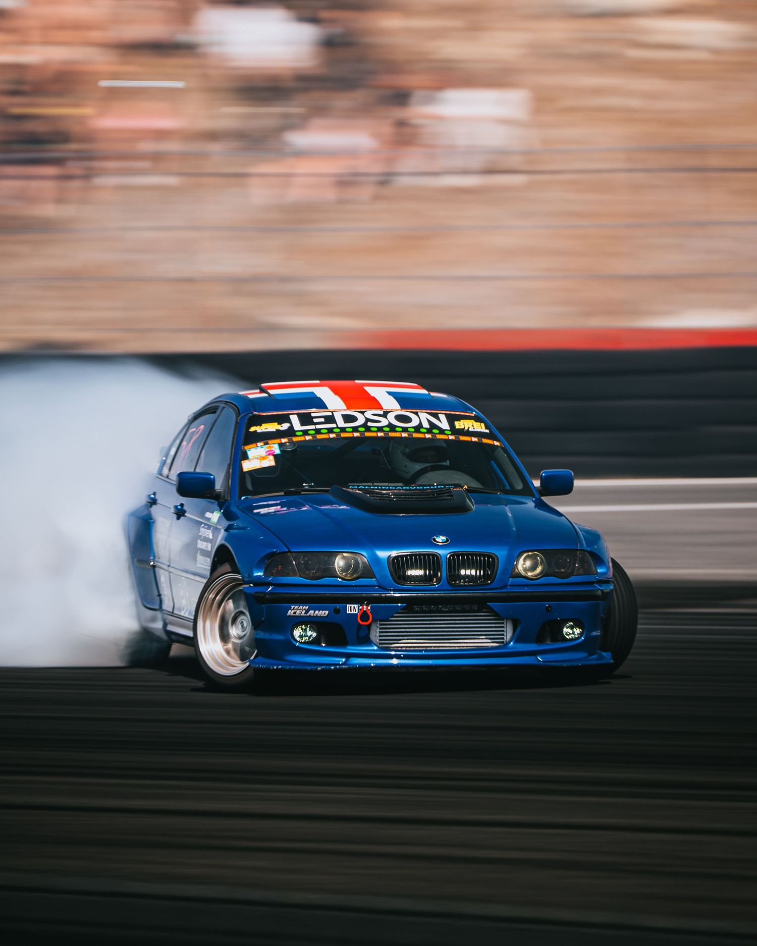 A blue drift car slides sideways creating smoke trails during a drift competition on a race track.