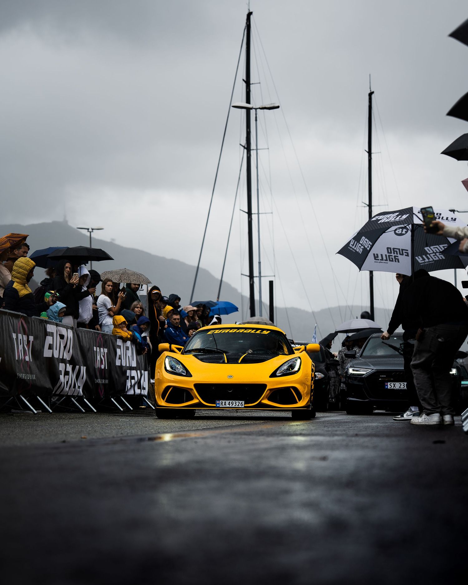 Bright yellow Lotus sports car navigating through spectators with umbrellas at a wet race event.