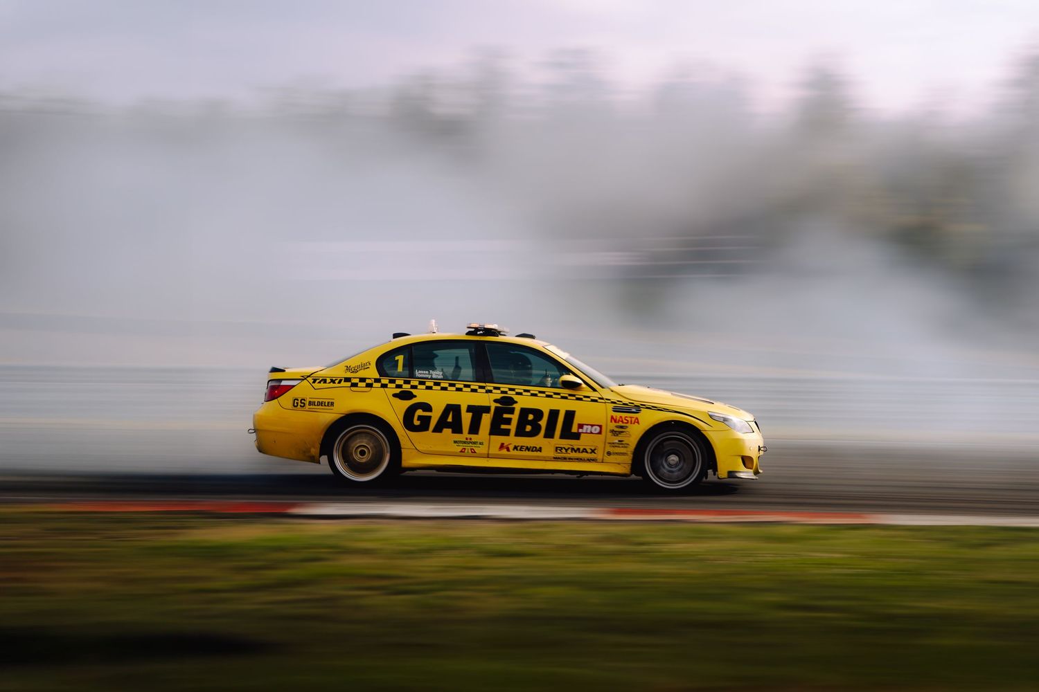 Yellow race car with Gatebil branding drifts on track creating smoke during motorsport competition.