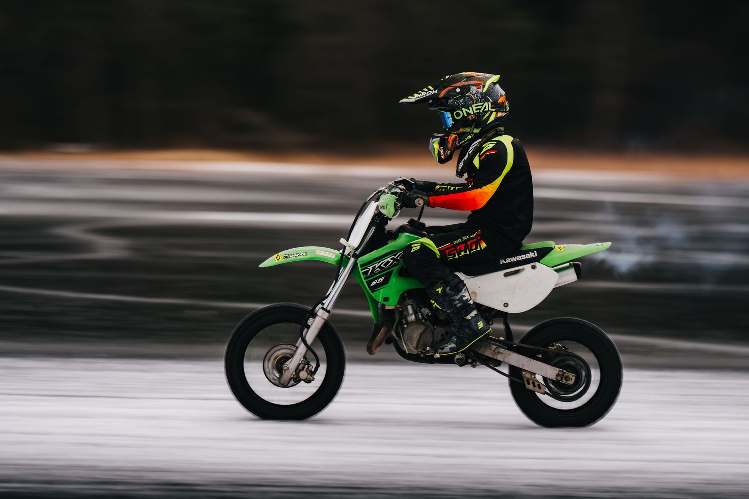 Motorcyclist performs high-speed ice racing on a frozen lake with a green dirt bike creating dramatic snow spray.