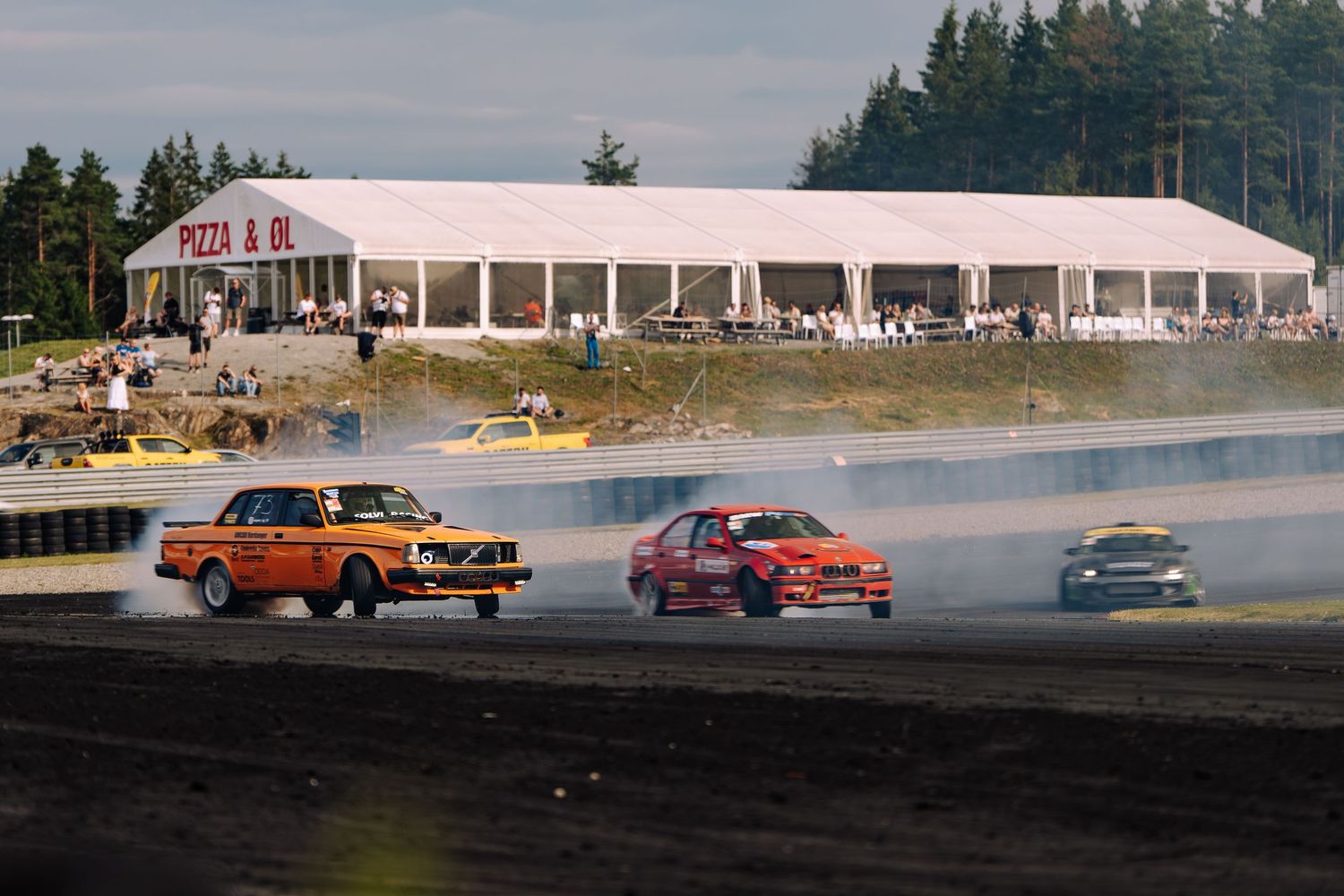 Race cars drift around a track corner leaving tire smoke trails during a competitive motorsport event.