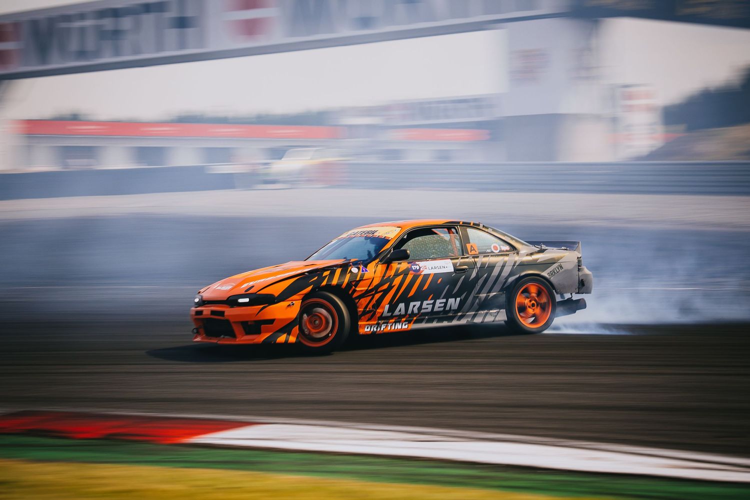 Orange drift car creates clouds of smoke while sliding sideways during a professional drifting competition.