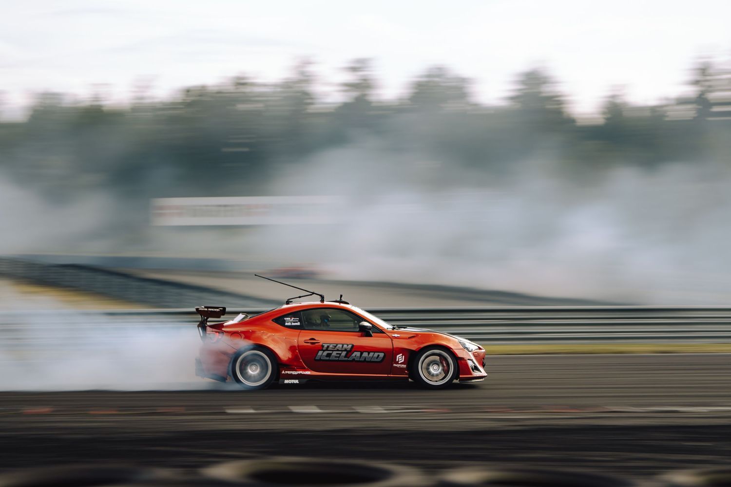 A red sports car drifts sideways on a race track creating dramatic tire smoke during a professional drift competition.