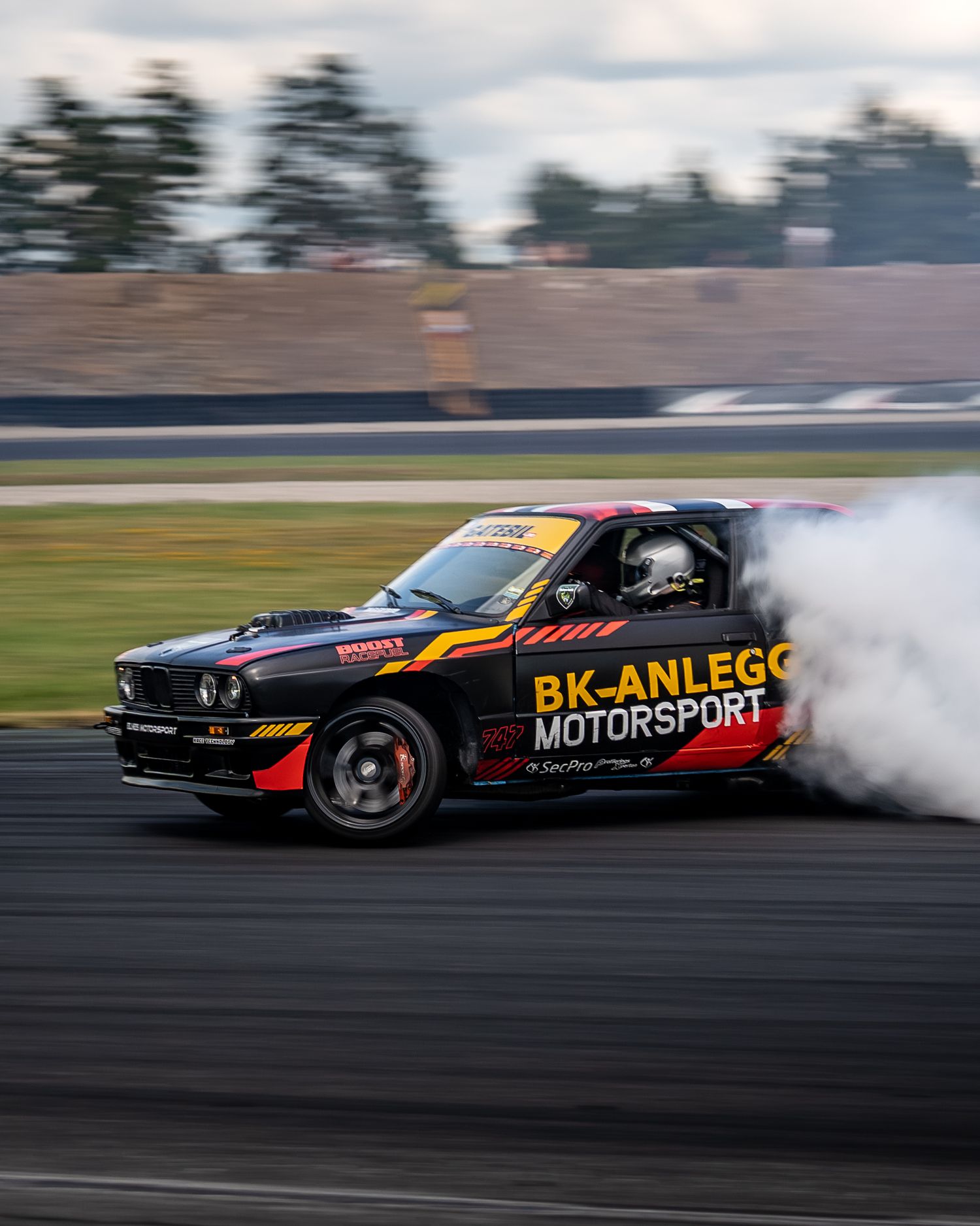 A black drift car with orange and red racing livery performs a smoky drift maneuver on a racetrack.