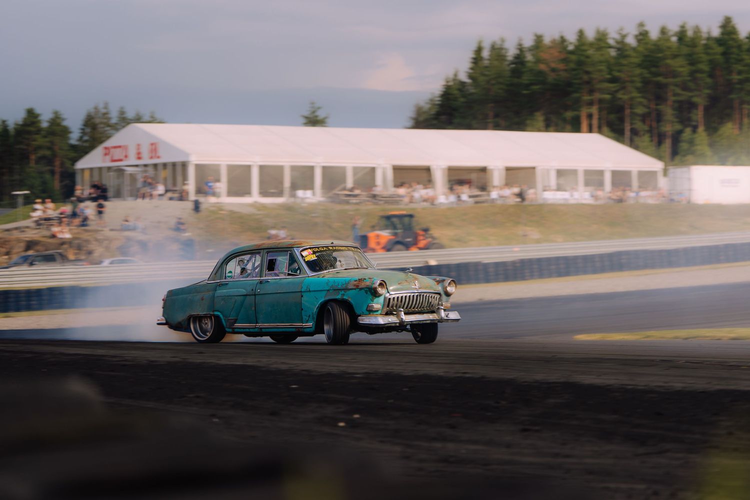 Vintage cars line up on a racetrack with evergreen trees and white buildings visible in the background.