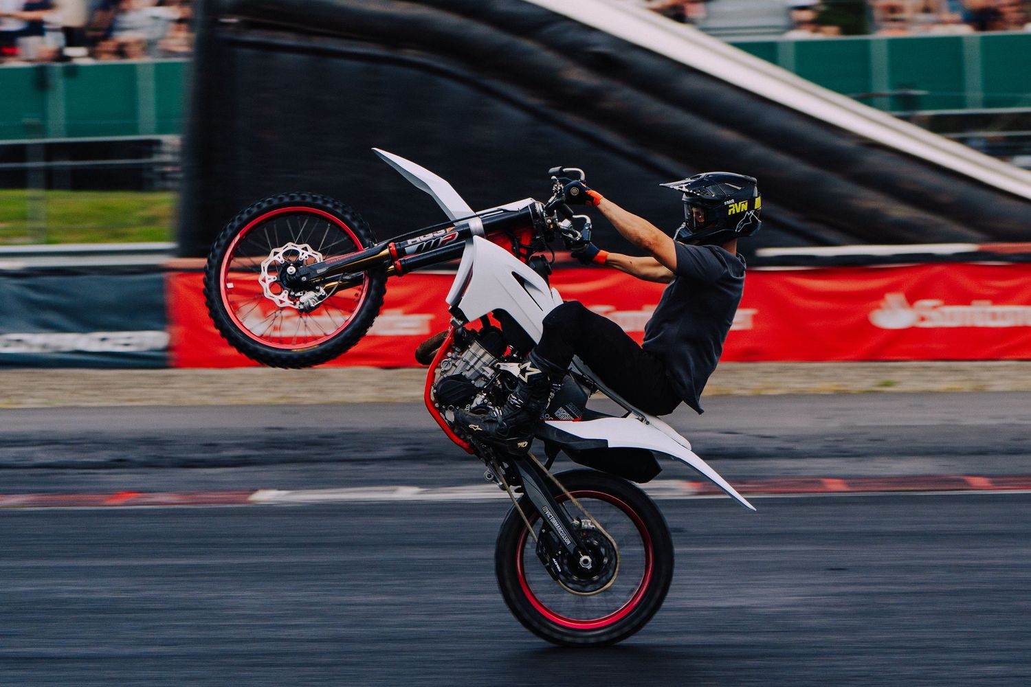 A motorcycle rider performs a wheelie stunt on a racetrack, lifting the front wheel high off the ground.