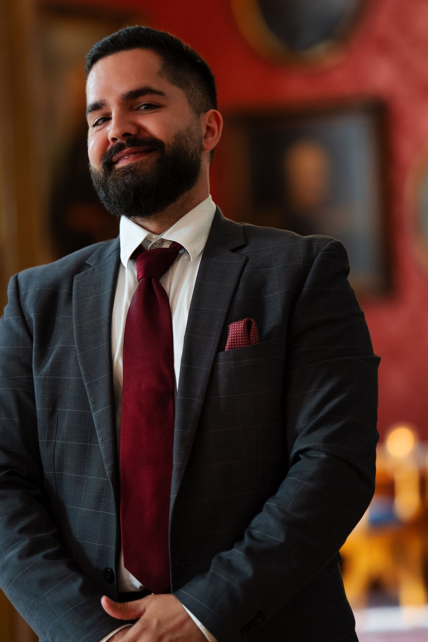 Professional in dark suit and burgundy tie poses against red wall at formal event.