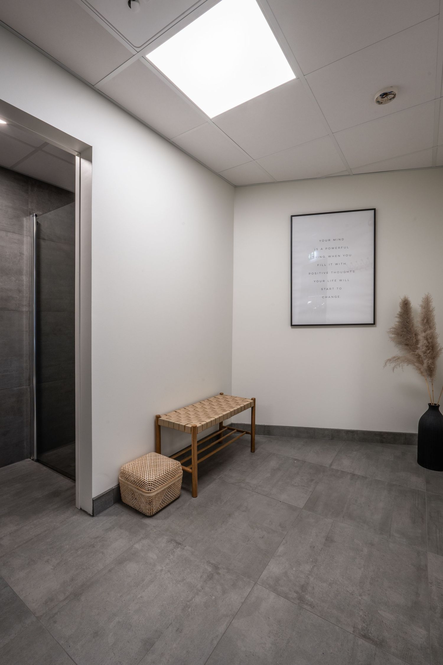 Waiting area with wooden bench, woven pouf and framed artwork near elevator entrance.