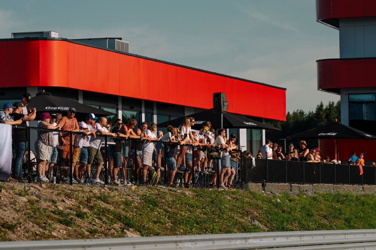 A crowd of spectators gathers outside a building with red architectural accents on a sunny day.