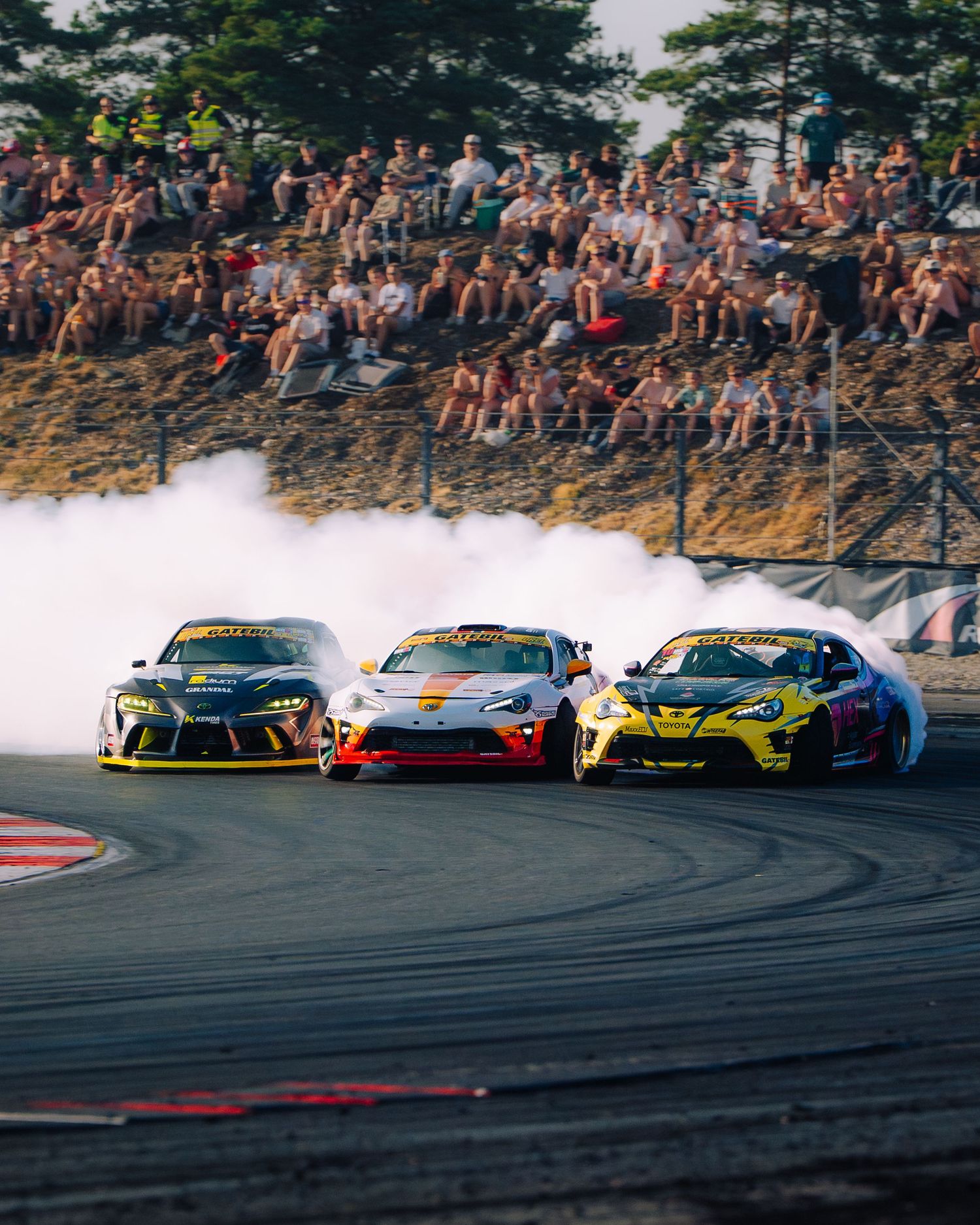 Three drift cars compete side by side creating smoke clouds during a professional racing event on a track.