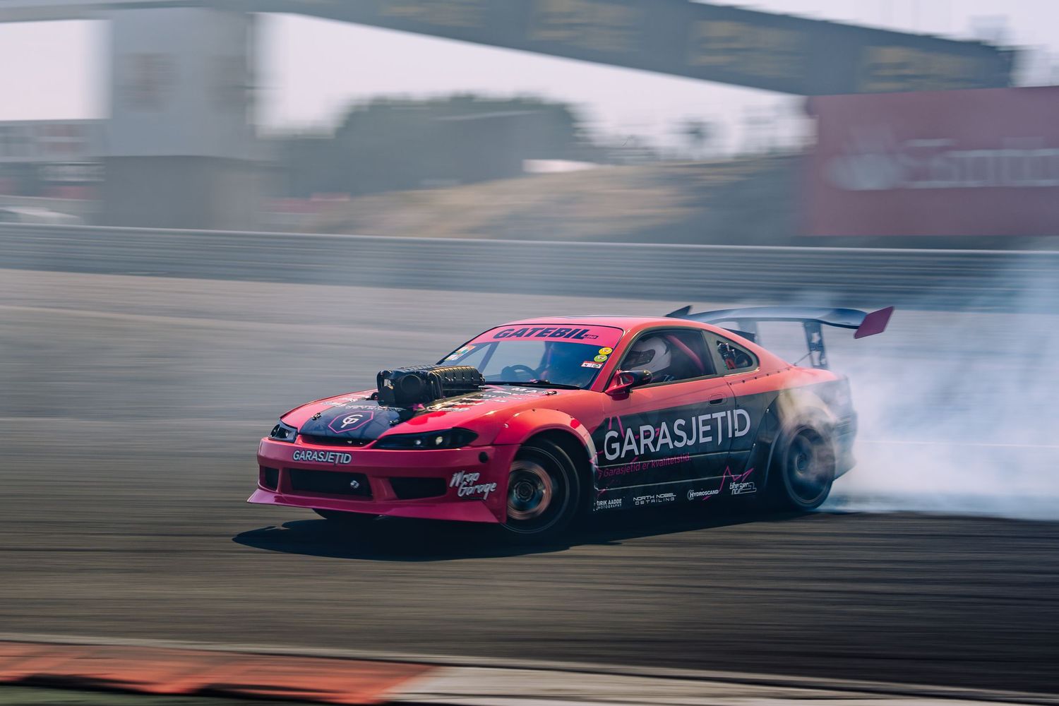 A red drift car slides sideways creating smoke while drifting on a race track during competition.