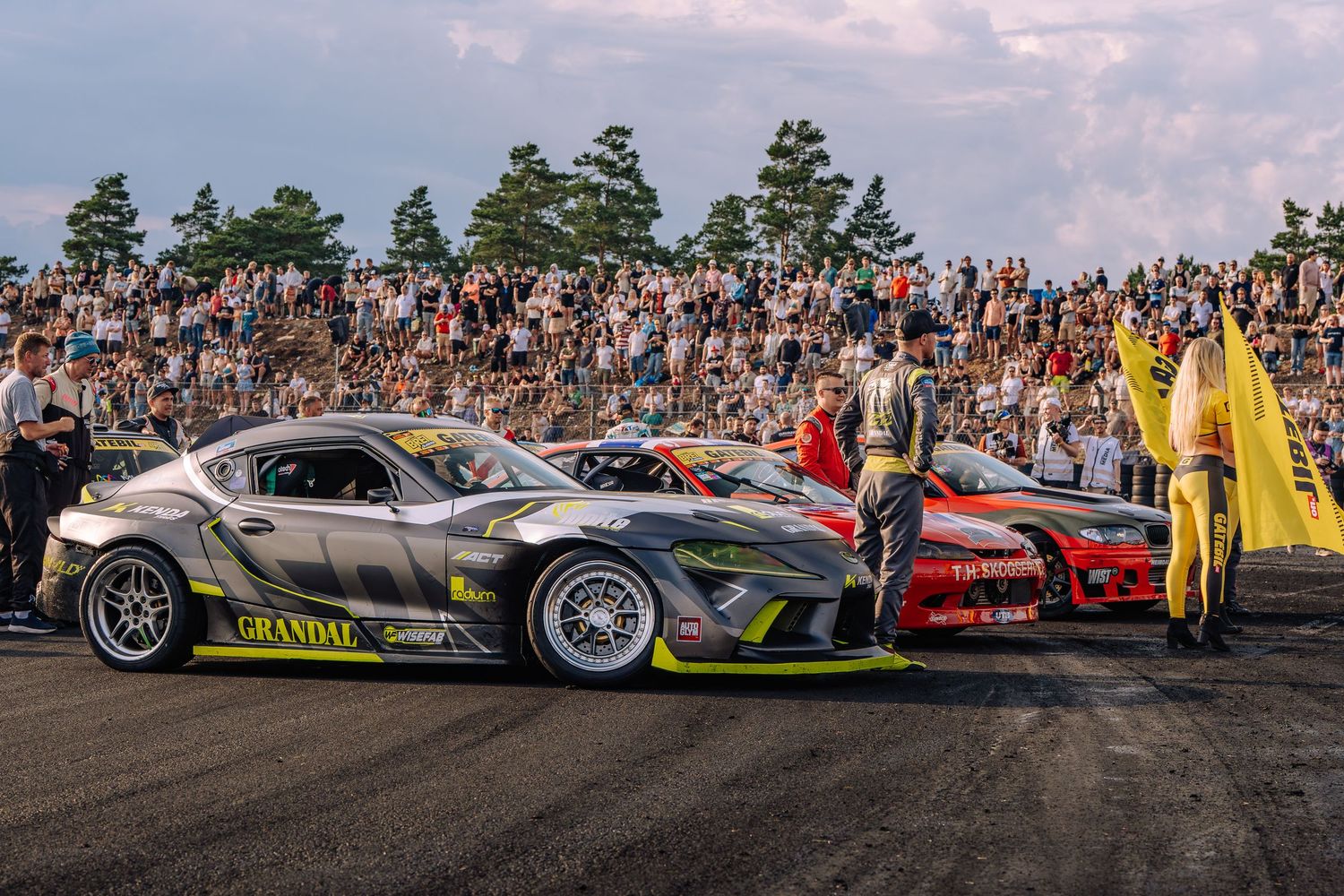 Custom sports cars line up at a drift event with spectators watching from a crowded hillside under cloudy skies.