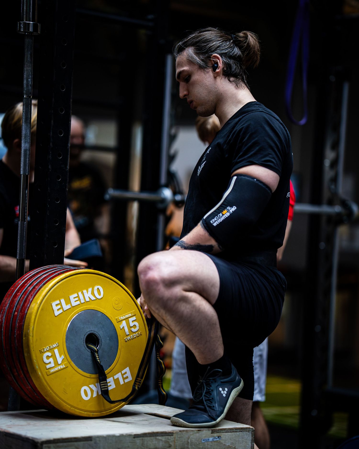 Athlete in black attire crouching next to a yellow Eleiko weightlifting plate in a gym setting.
