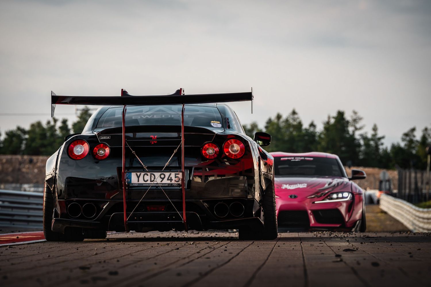 A line of red and black sports cars parked on a racetrack with dramatic cloudy skies in the background.