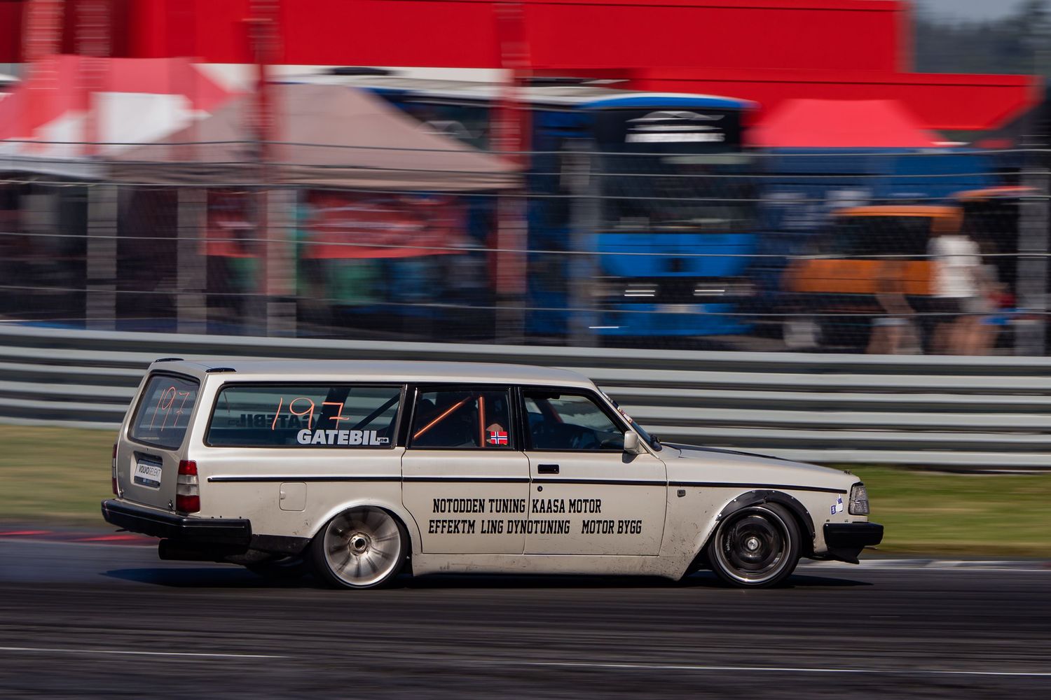 White vintage race car speeds around track during competition event with red and blue barriers visible.