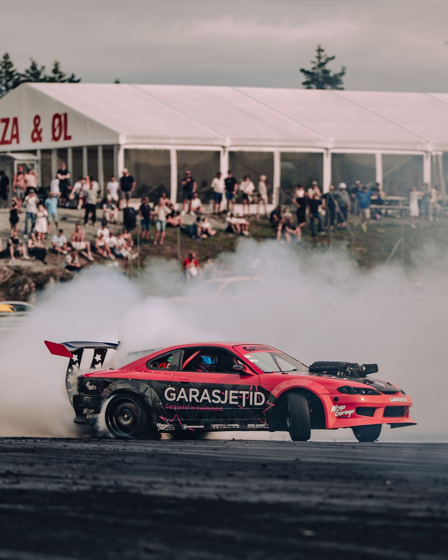 A red drift car performs a smoky burnout on a track while spectators watch from behind barriers at an outdoor event.