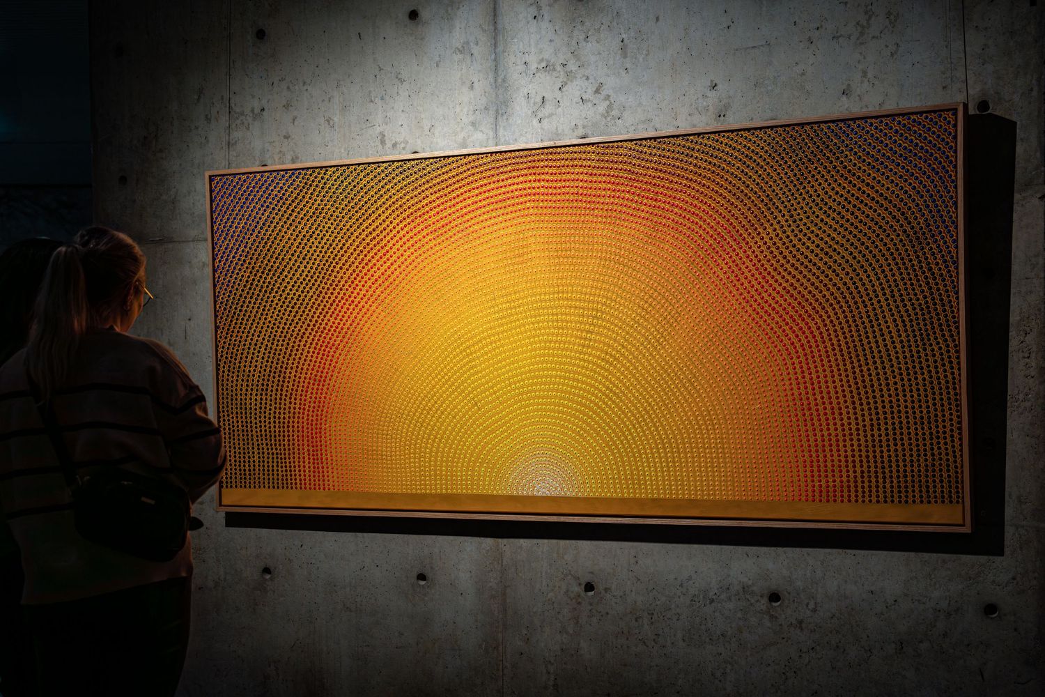 A visitor observes a large orange geometric artwork with radiating lines mounted on concrete wall.