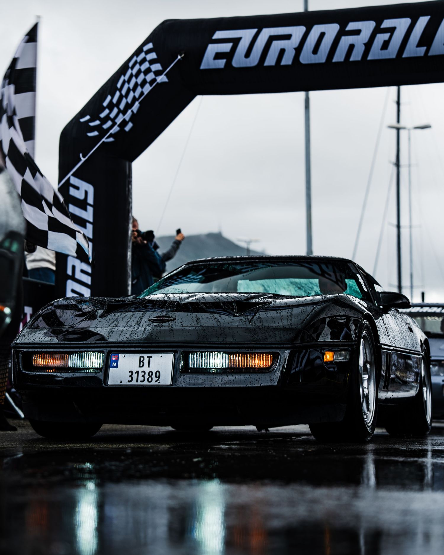 Black vintage sports car passing under Eurorally arch on wet race track during rainy day.