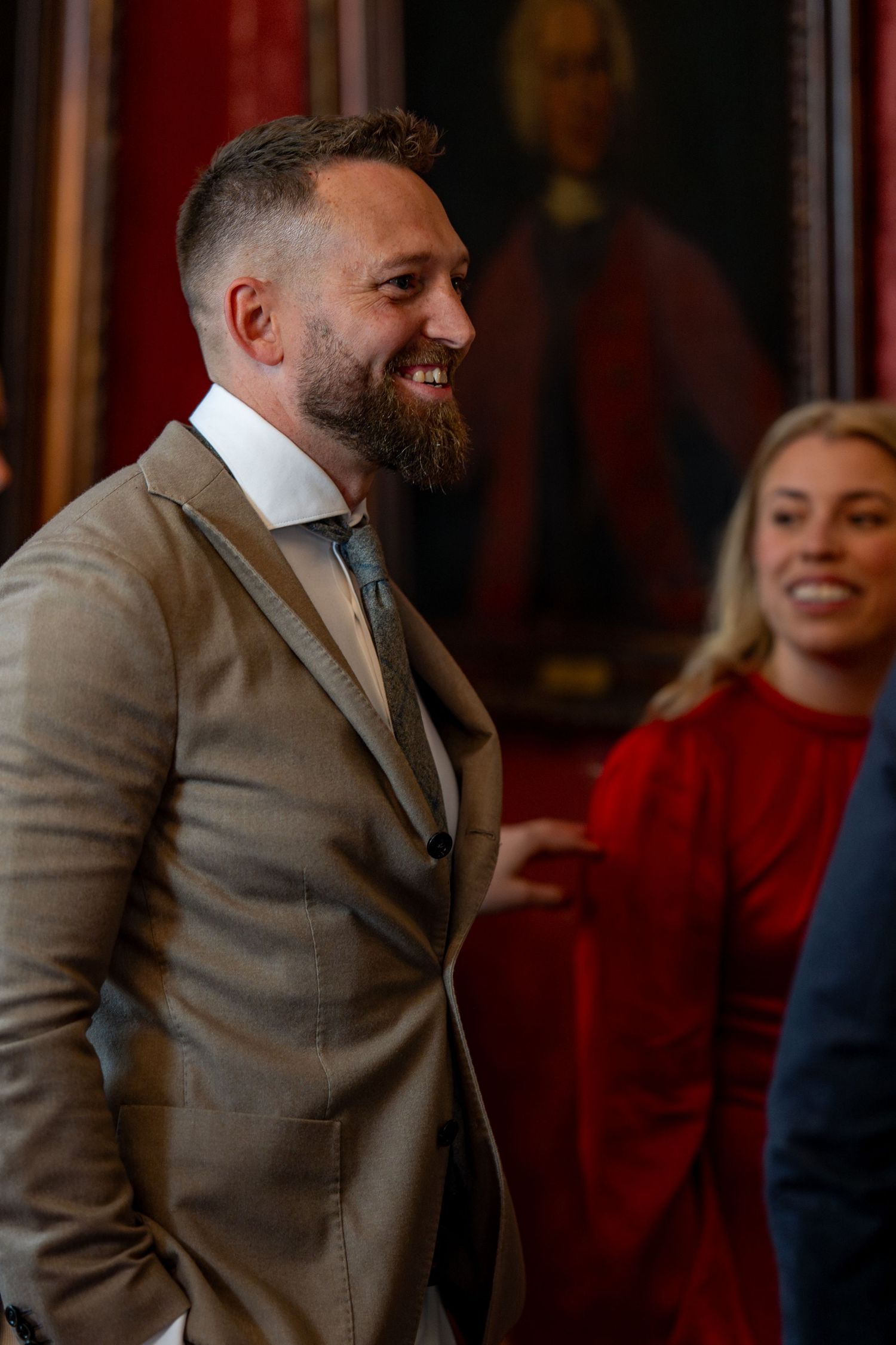 Well-dressed man in gray suit smiles during formal gathering with red wall background.