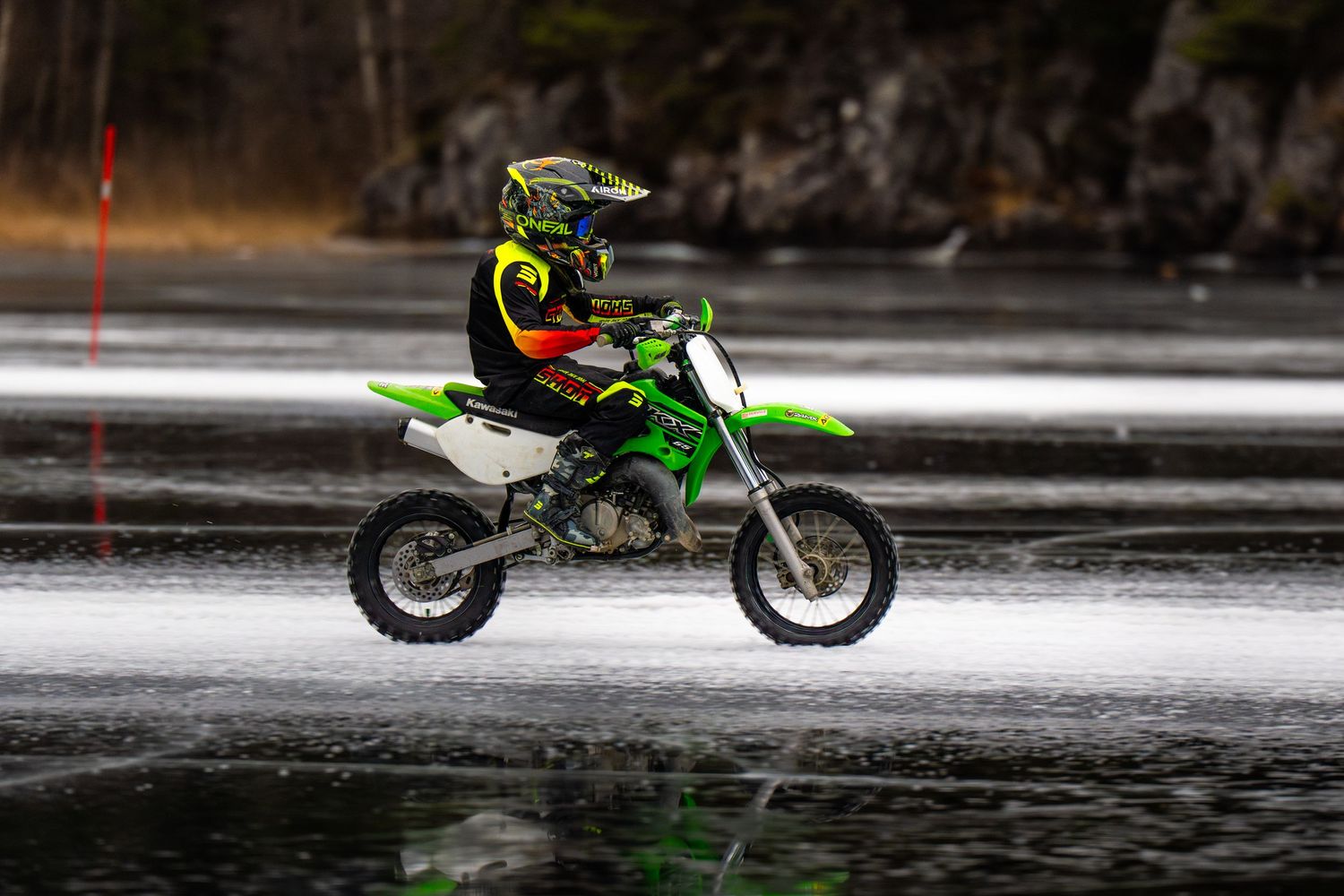 A dirt bike rider performs a spectacular water skimming stunt across a lake, creating a dramatic splash trail behind.
