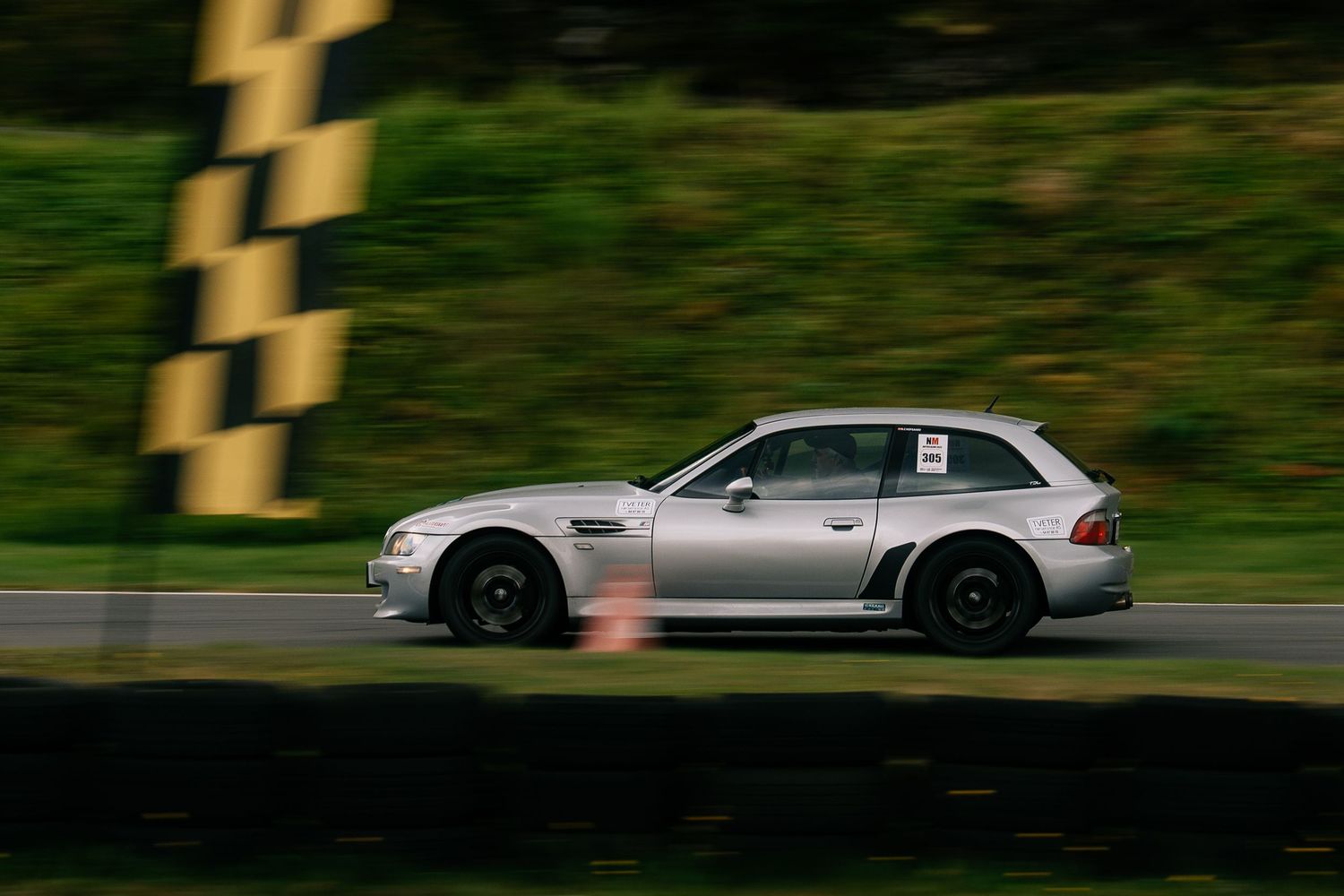 Silver BMW Z3 roadster speeds along a winding race track with green foliage in the background.