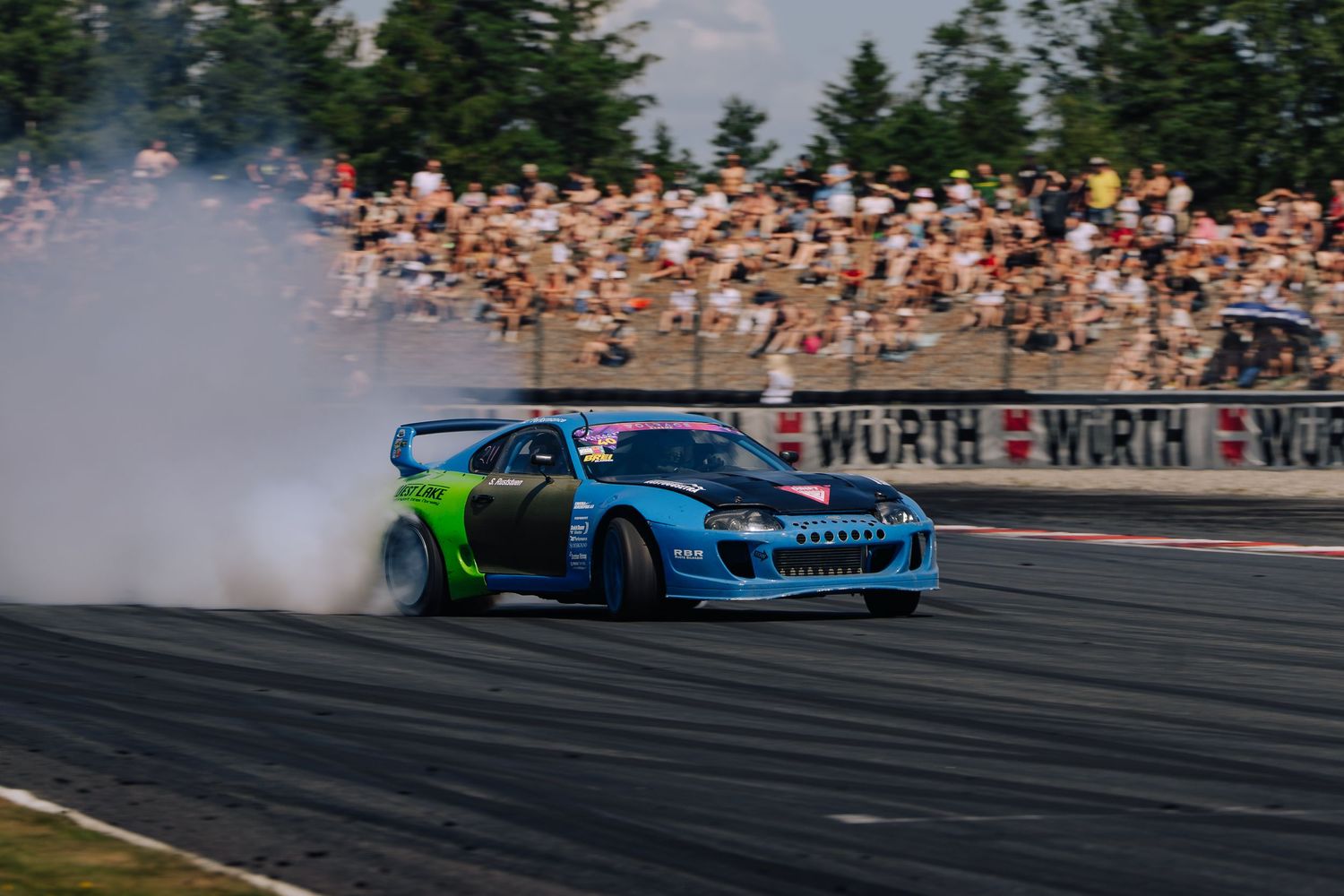 A blue drift car performs a smoky burnout on a racetrack in front of a large crowd of spectators.