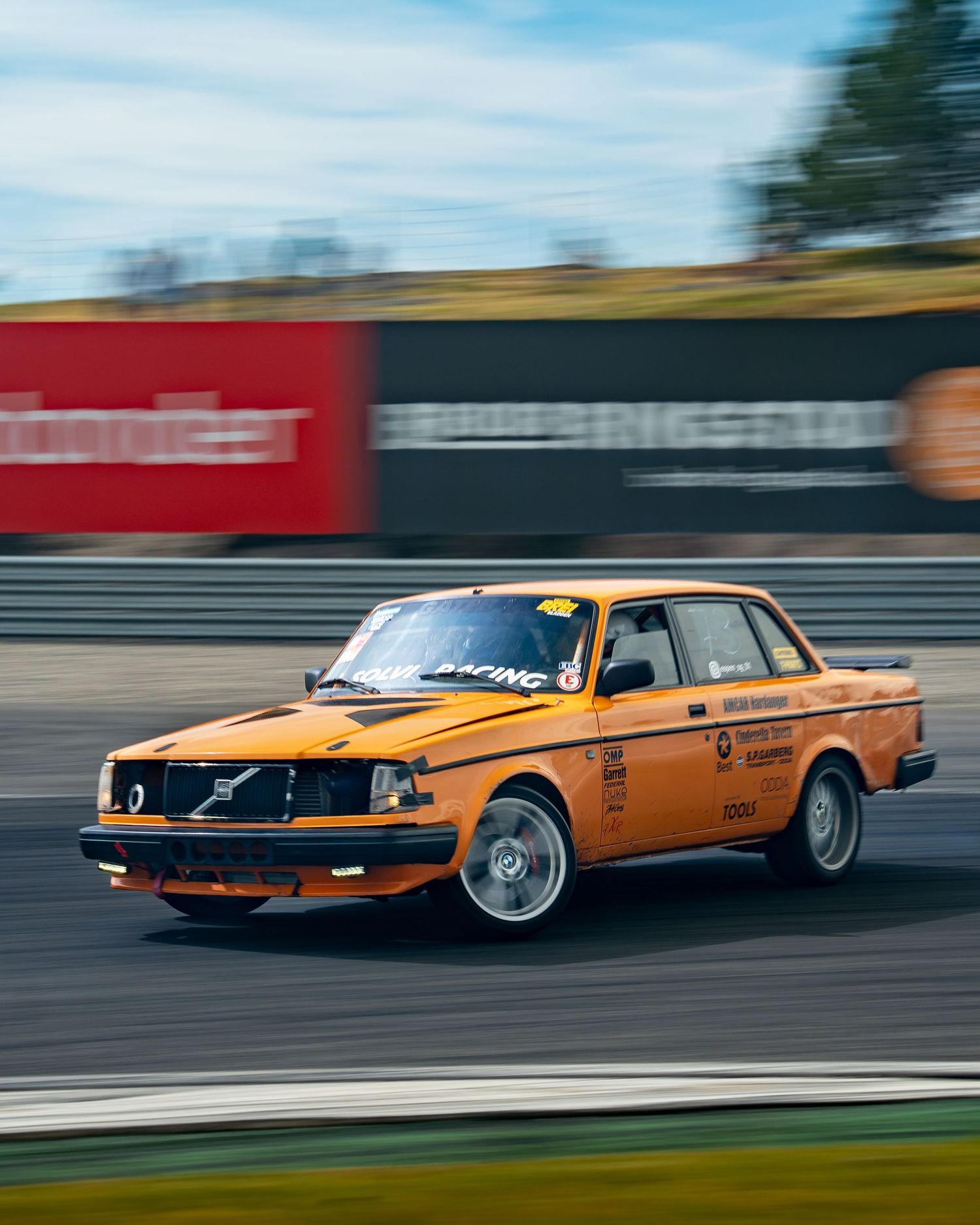 An orange Volvo 240 race car drifting on a racetrack with motion blur and advertising barriers in the background.