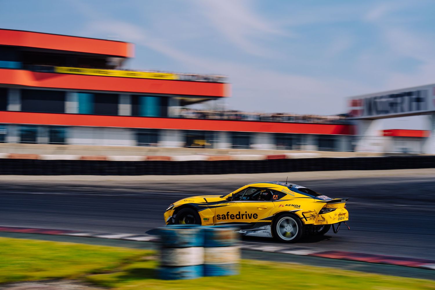 Yellow racing car speeds around a racetrack with red and white barriers visible in the background.