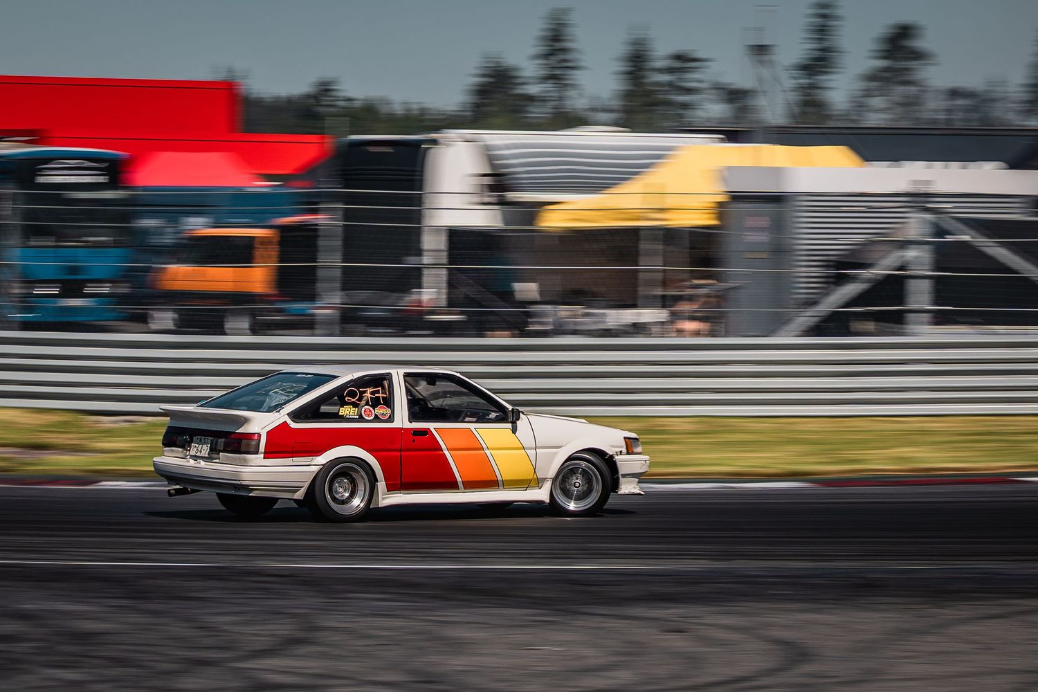 Colorful vintage cars racing down a drag strip with motion blur effect and spectators in the background.
