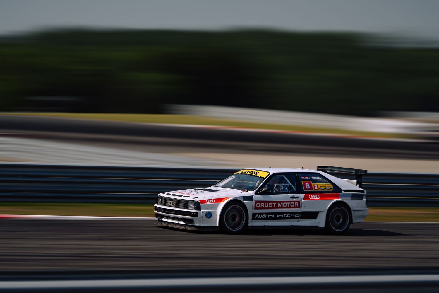 A white Mitsubishi race car speeds along a racetrack with motion blur effect in the background.