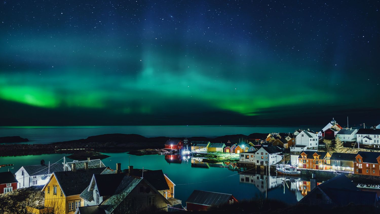 Green northern lights dancing above a colorful coastal town with houses reflected in harbor waters.