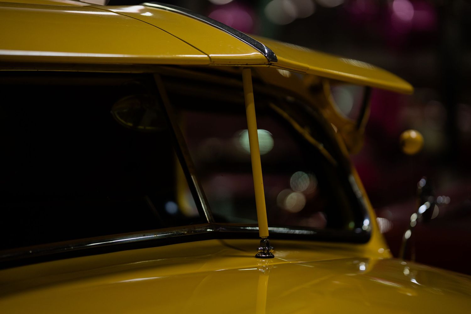 Yellow classic car detail showing antenna and windshield at indoor car show.
