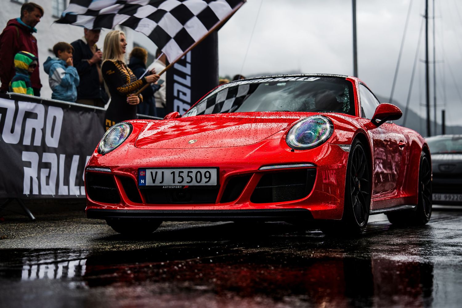 Red Porsche 911 crossing finish line at Eurorally event in rainy conditions.